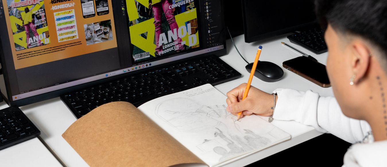 Graphic Design student at SBC sketches in a notebook while sitting at a desk with three computer monitors displaying colorful graphic designs, suggesting a creative workspace.