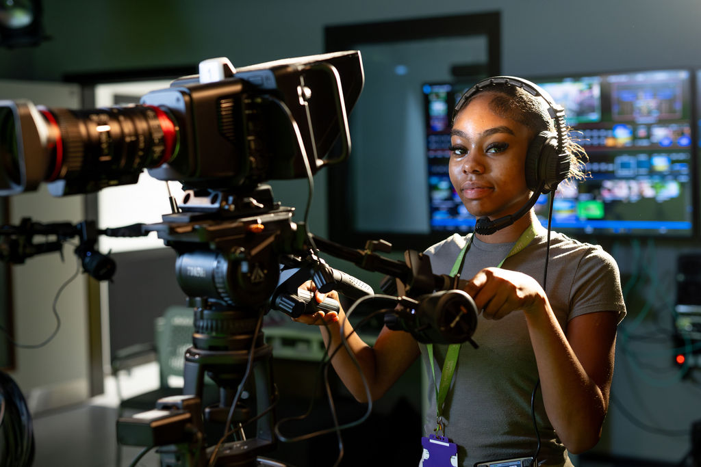 Confident LSBTC student wearing headphones operates a professional video camera in a broadcast studio, with glowing monitors behind her.