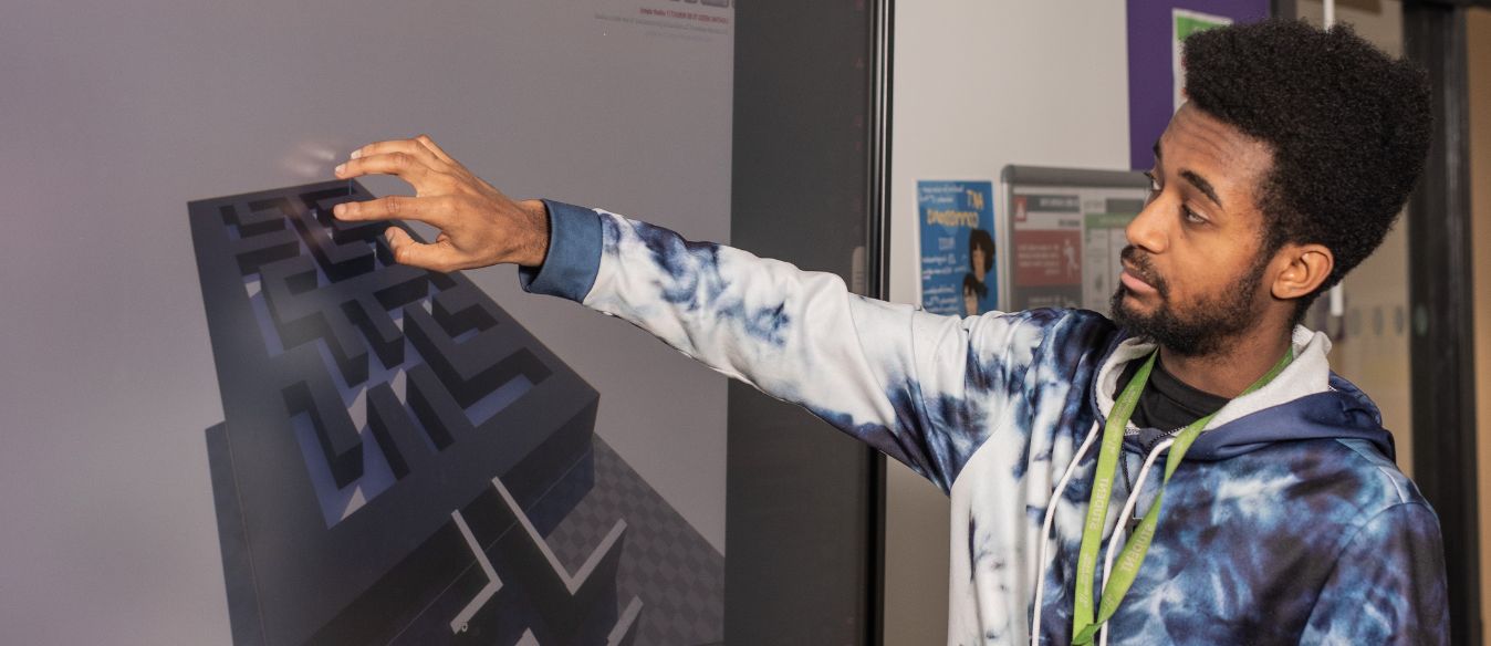 A LSBTC games design student gestures towards a large screen with a 3d model while teaching in a classroom setting.