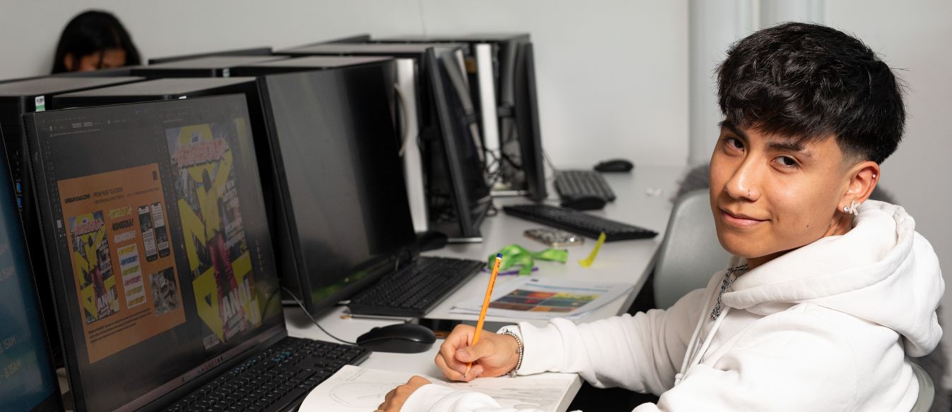 A LSBTC graphic design student smiling to the camera whilst sitting in front of a computer with graphics on the screen and holding a pencil