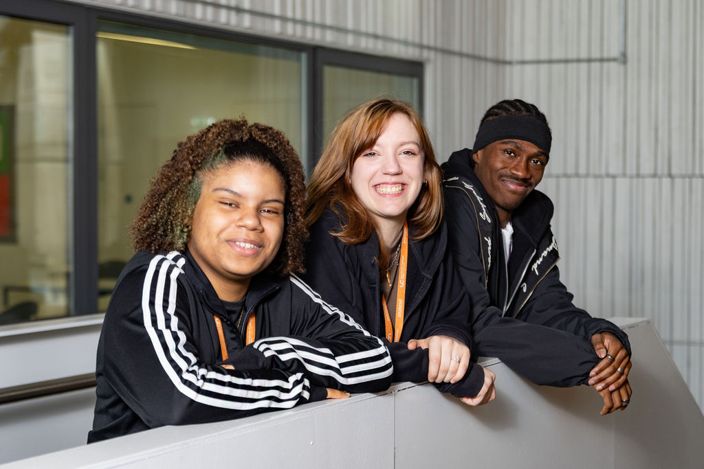 Three diverse young adults in black athletic jackets smile and lean over a railing indoors, conveying friendship and upbeat energy.
